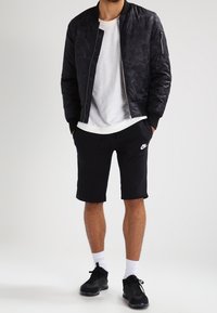 Svart bomberjacka med ett subtilt mönster, vit t-shirt under, svarta shorts och svarta sportskor med vita strumpor.