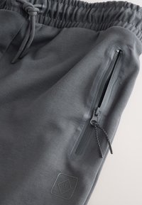 Gray jogger shorts made of smooth fabric with an elastic waistband, featuring a side zip pocket and a logo patch near the hem.