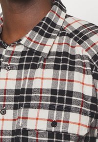 Camicia di flanella con un motivo a quadri nero, bianco e rosso; presenta un colletto e una chiusura con bottoni; due tasche sul petto.