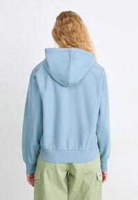 Sudadera azul claro con capucha, puños y dobladillo acanalados. Tela suave, corte ligeramente relajado. Combinada con pantalones cargo verdes con bolsillos.