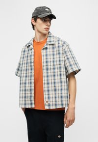 Νέος άνδρας που φοράει μπλε και άσπρο πουκάμισο με κοντά μανίκια και καρό σχέδιο πάνω από πορτοκαλί μπλουζάκι, μαύρο παντελόνι και μαύρο καπέλο, κοιτάζει προς τα αριστερά.