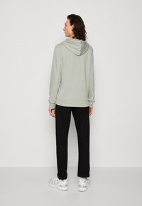 Felpa verde chiaro con cappuccio regolabile, abbinata a pantaloni neri e sneakers bianche. Il tessuto sembra morbido e comodo.