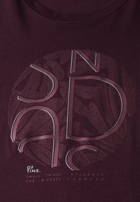 Camiseta burdeos con un gráfico circular que presenta texto estilizado y patrones de líneas intrincados en colores apagados. Mangas cortas y cuello redondo.