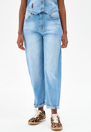 Persoon draagt lichtblauwe high-waist spijkerbroek met omgeslagen pijpen en sneakers met luipaardprint en witte veters, staand tegen een witte achtergrond.