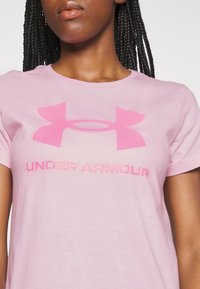 Ljusrosa t-shirt med korta ärmar och en stor rosa Under Armour-logga samt text i en något mörkare nyans, tillverkad av mjukt bomullstyg.