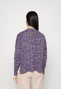 Femme aux longs cheveux noirs portant un pull à imprimé animalier violet et un pantalon beige clair, vue de dos sur un fond blanc uni.
