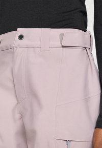 Világos rózsaszín nadrág, lapos derékpánttal, oldalzsebekkel, egy cipzáras zsebbel és sima, texturált anyagból.