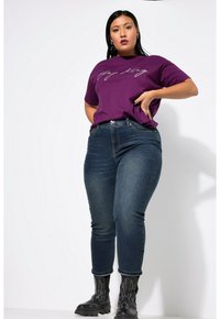 Camiseta corta de manga en color púrpura con script blanco, combinada con jeans ajustados de color azul oscuro y botas negras hasta el tobillo con costuras decorativas.