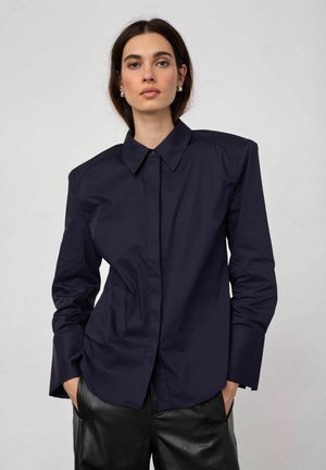 Femme aux cheveux foncés portant une chemise bleu marine foncé à boutons avec épaules rembourrées et un pantalon en cuir noir sur un fond clair uni.