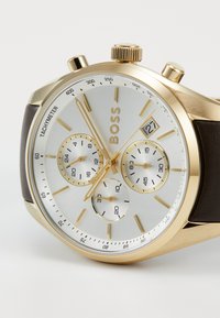 Reloj cronógrafo en tono dorado con esfera plateada, correa de cuero marrón, indicador de fecha, tres subesferas y marcas de taquímetro en el bisel.