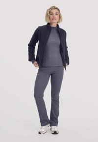 Frau steht in einer dunklen Fleece-Jacke über einem grauen Zip-Up-Top, passenden Hosen und weißen Sneakers vor einem schlichten hellen Hintergrund.