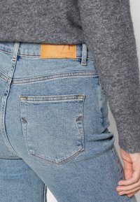 Jeans in denim azzurro chiaro con cuciture visibili e dettagli sulla tasca posteriore, con un'etichetta in pelle beige che recita "SELECTED". Pullover grigio parzialmente visibile.