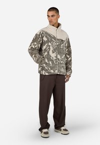 Beżowa i szara bluza polarowa z abstrakcyjnymi wzorami, wysoki kołnierz i pełny zamek. Połączona z ciemnobrązowymi spodniami i kremowymi sneakersami.