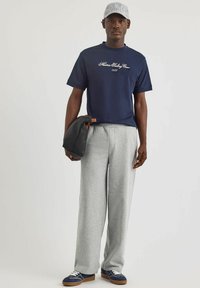 T-shirt blu con testo ricamato bianco, pantaloni felpati grigi e scarpe da ginnastica blu con lacci bianchi. Borsa nera in mano, indossando un cappellino grigio a righe.