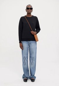 Felpa nera con polsini a coste, jeans in denim blu chiaro con un motivo di logo scuro, scarpe nere con tacco basso e una borsa intrecciata marrone.