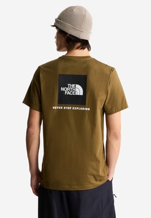 Mężczyzna ubrany w oliwkowo-zieloną koszulkę The North Face z logo i napisem „Never Stop Exploring” na plecach, beżową dzianą czapkę oraz ciemne spodnie.