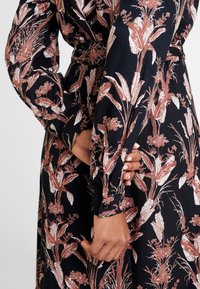 Gros plan d'une personne portant une robe noire avec des motifs floraux rouille et blancs, des boutons aux poignets et une main qui tient légèrement le tissu.