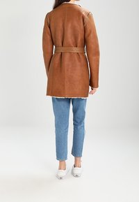 Abrigo de cuero marrón con cinturón en la cintura, con una textura suave y detalles en los hombros, combinado con vaqueros azul claro y zapatillas blancas.