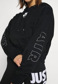 Sudadera negra de Nike con un logo en el frente y texto grande en blanco en las mangas. La tela es suave, con un ajuste relajado y capucha con cordón.