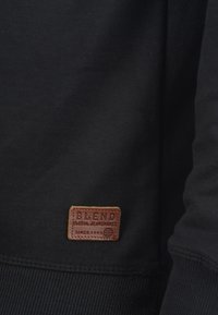 Tissu de sweat noir avec des poignets côtelés et une patch en cuir marron portant l'inscription "BLEND GLOBAL JEANMAKER" en bas à gauche.