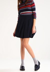 Falda plisada azul marino combinada con un suéter de cuello alto a rayas rojas y azules. La modelo lleva calcetines grises y mocasines negros. Tela suave, corte clásico.