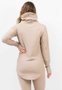 Beige lange mouw top met een hoge hals, geribbelde textuur en een gebogen zoom. Het is gemaakt van een rekbare, gladde stof voor flexibiliteit.