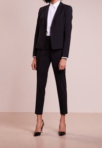 Veste de costume noire cintrée avec deux boutons, associée à un pantalon slim noir et une chemise blanche à col; des escarpins noirs en vernis complètent le look.