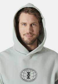 Světle zelená mikina s kapucí, která má přední kapsu a černý kruhový design loga. Tkanina vypadá jemně s hladkým povrchem. Kapuce je prostorná.