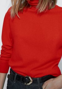 Pull à col roulé rouge en tricot avec des manches longues. Assorti à une ceinture en cuir noir et un jean en denim foncé, mettant en avant un design épuré.