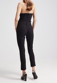 Mono negro sin tirantes con un diseño ajustado, cierre de cremallera en la parte posterior y pantalones de corte slim. Se combinan con tacones gruesos negros.