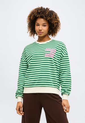 Tânără cu păr creț purtând un pulover verde cu dungi albe cu text roz „Colour Fly Rebel” și pantaloni maro, stând pe un fundal alb.