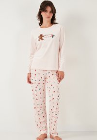 Pitkähihaiset vaaleanpunaiset pyjaman paita, jossa on piparkakkumiehen grafiikka ja teksti, yhdistettynä vaaleanpunaisiin housuihin, joissa on värikkäitä kukka- ja tähtikuvioita.