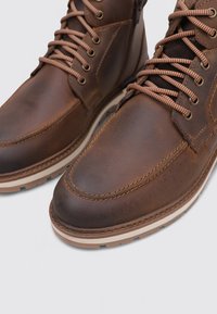 Botas de tobillo de cuero marrón con punta redonda, costuras en contraste y suelas de goma beige. Atadas con cordones marrones oscuros y beige.