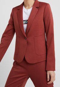 Blazer bordeaux à coupe ajustée, fermeture à un bouton, et deux poches avant. Texture lisse, col à revers, porté avec un t-shirt blanc.