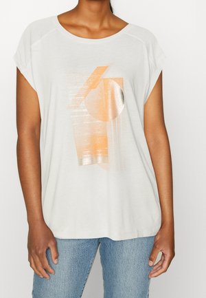 Femme portant un haut blanc à manches courtes avec des formes géométriques abstraites pêche et argent, associé à un jean bleu clair.