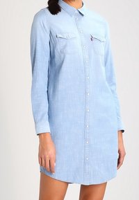 Světle modré džínové šaty košového stylu s dlouhými rukávy, prošitým předním zapínáním na knoflíky, dvěma náprsními kapsami, zaobleným lemem a volným střihem.