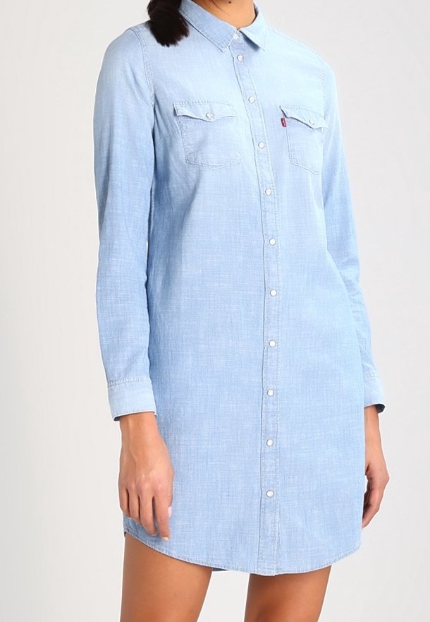 Světle modré džínové šaty košového stylu s dlouhými rukávy, prošitým předním zapínáním na knoflíky, dvěma náprsními kapsami, zaobleným lemem a volným střihem.