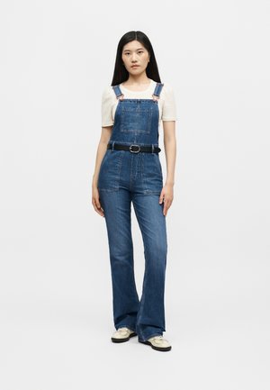 Vrouw staat, draagt blauwe denim overalls met een zwarte riem, wit top met korte mouwen en crèmekleurige schoenen tegen een effen witte achtergrond.