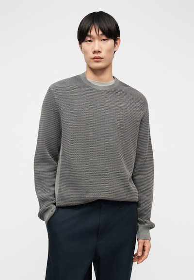 Jeune homme portant un pull à col rond gris texturé et un pantalon bleu marine foncé, debout avec une main dans la poche devant un fond uni.