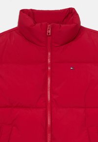 Giacca a vento rossa con colletto alto e zip frontale. Presenta un piccolo logo sul lato sinistro, texture trapuntata e tasca laterale.