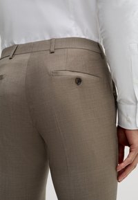Zbliżenie osoby noszącej beżowe spodnie garniturowe z zapiętymi na guziki tylnymi kieszeniami oraz białą koszulę na długim rękawie, z dłonią spoczywającą wzdłuż boku.