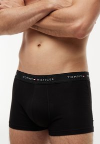 Αρσενικός κορμός με μαύρα εσώρουχα Tommy Hilfiger, χέρια σταυρωμένα πάνω στο γυμνό στήθος, με λευκό φόντο.