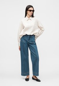 Γυναίκα με μακριά σκούρα μαλλιά, φορώντας γυαλιά ηλίου, χαλαρό μπεζ πουκάμισο με κουμπιά, μπλε τζιν παντελόνι με φαρδιές μπατζάκια και μπροστινές τσέπες και μαύρα φλατ παπούτσια, όρθια.