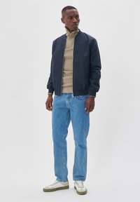 Marineblauwe bomberjack over een beige sweater met rits, gecombineerd met lichtblauwe jeans en witte sneakers met olijfgroene zolen.