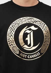 Μαύρο πουκάμισο με χρυσαφί κυκλικό λογότυπο που απεικονίζει δύο διαπλεκόμενα κεφάλια φιδιών και στιλιζαρισμένα γράμματα "CJ" πάνω από το κείμενο "JUST CAVALLI".