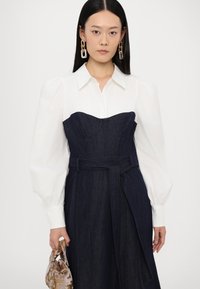Denim jurk met een gestructureerde corresponderende bovenkant, een witte blouse met lange mouwen en een ceintuur om de taille. Bevat puff mouwen en een handtas met bloemmotief.
