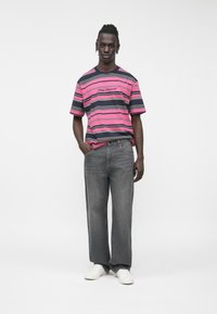 Maglietta a righe rosa e blu navy con logo ricamato, abbinata a jeans grigi a gamba larga e sneakers bianche, su uno sfondo semplice.