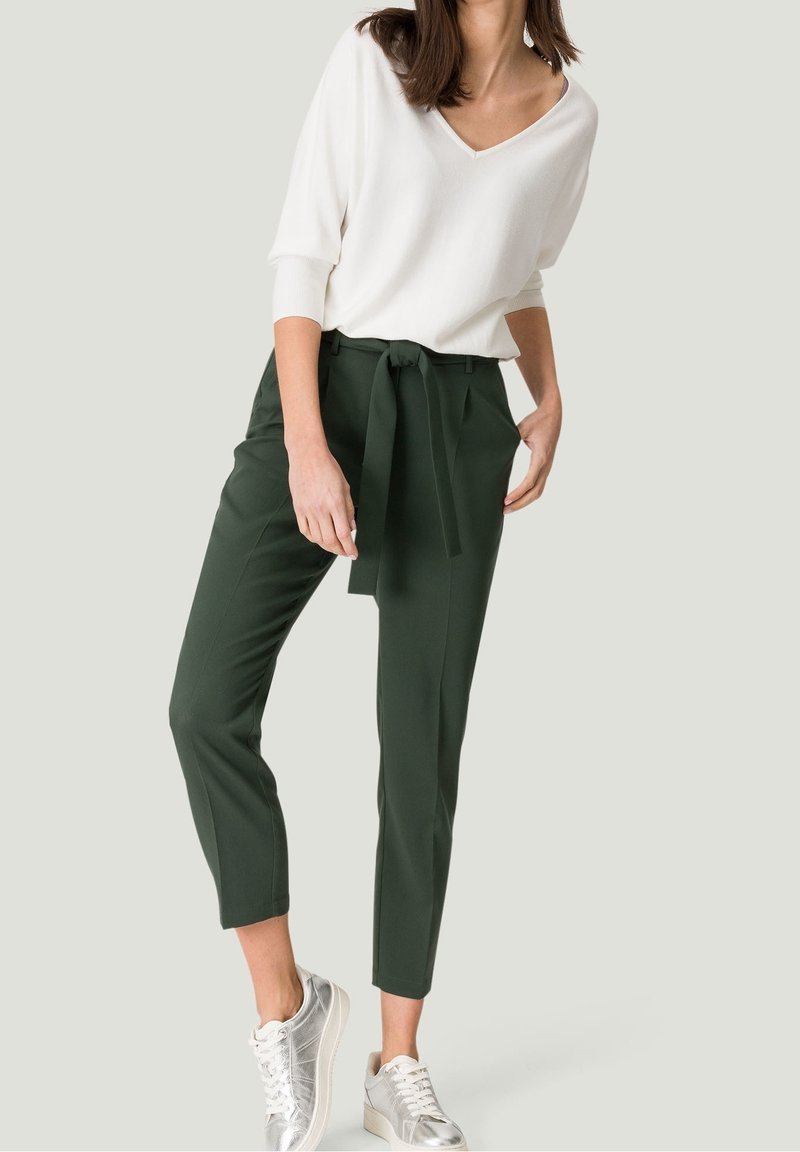 Pantalons taillés verts avec une ceinture à nouer, associés à un haut blanc à col en V. Le modèle porte des baskets argentées avec des détails texturés.