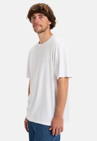 Bílá bavlněná tričko s krátkým rukávem, kulatým výstřihem a jemným logem značky na hrudi. V kombinaci s modrými džíny.