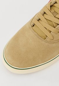 Hellbraune Wildledersneaker mit runder Spitze, strukturierter Oberfläche, beigen Schnürsenkeln und einer weißen Zwischensohle, die durch einen grünen Streifen akzentuiert ist.